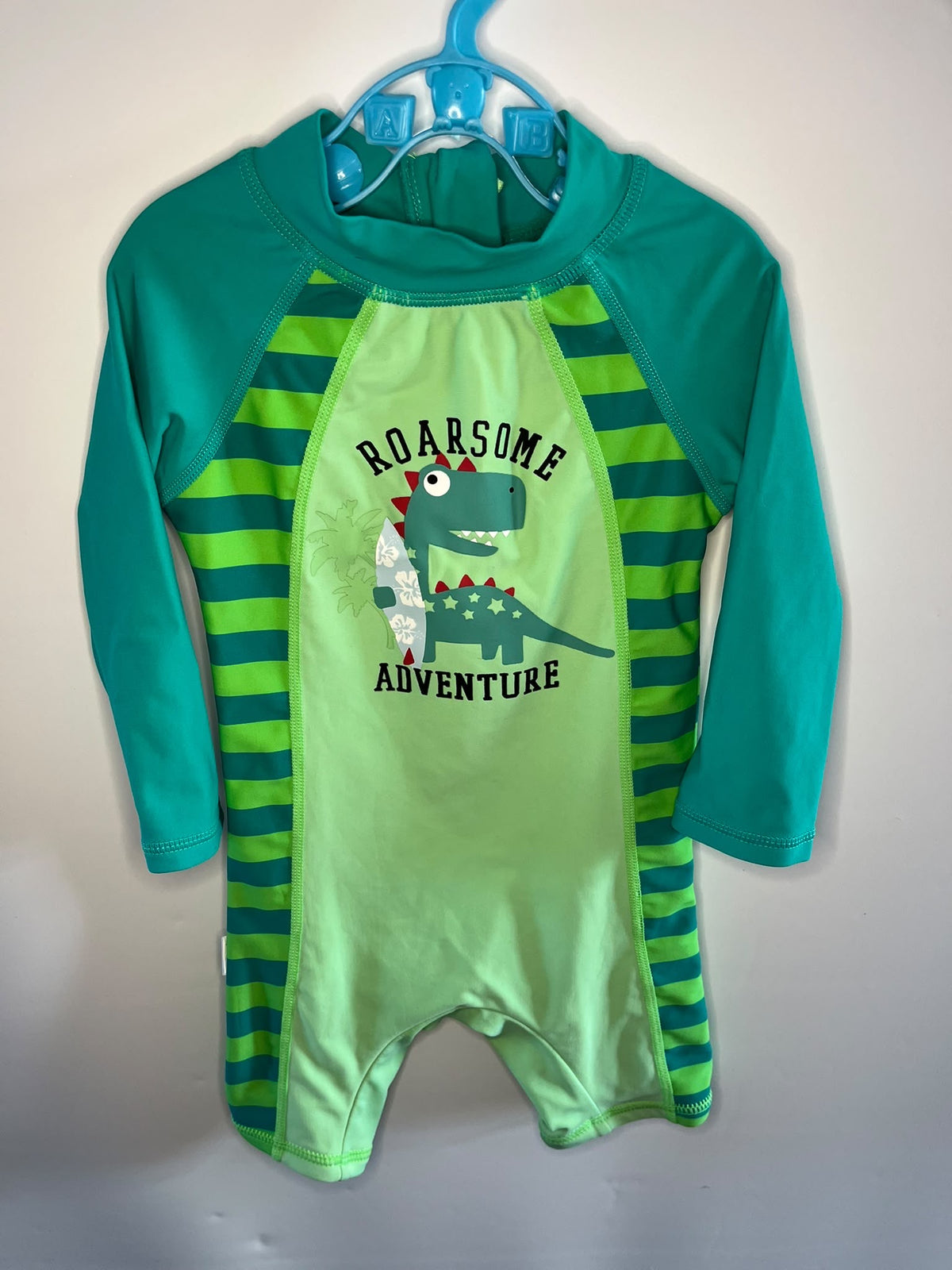A toddler's long-sleeve one-piece swimsuit hanging on a blue plastic hanger against a plain light gray wall. The swimsuit features a bright lime green center panel with a cartoon dinosaur graphic and the words 'ROARSOME ADVENTURE' in black, teal green raglan sleeves, and dark green and teal horizontal stripes on the sides.