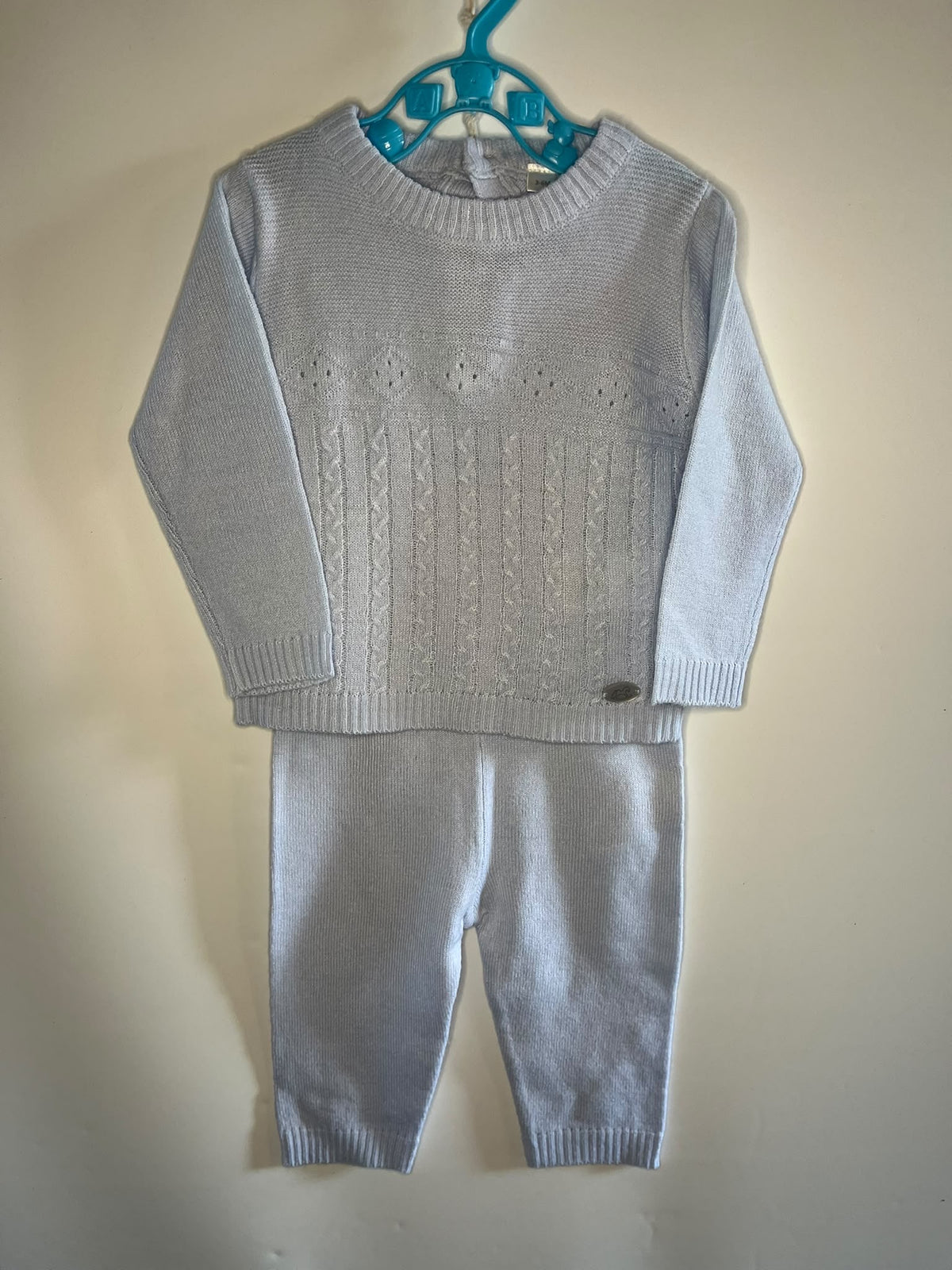 A baby or toddler knit two-piece outfit consisting of a light blue long-sleeve sweater and matching knit pants, hanging on a turquoise plastic hanger against a plain beige wall.