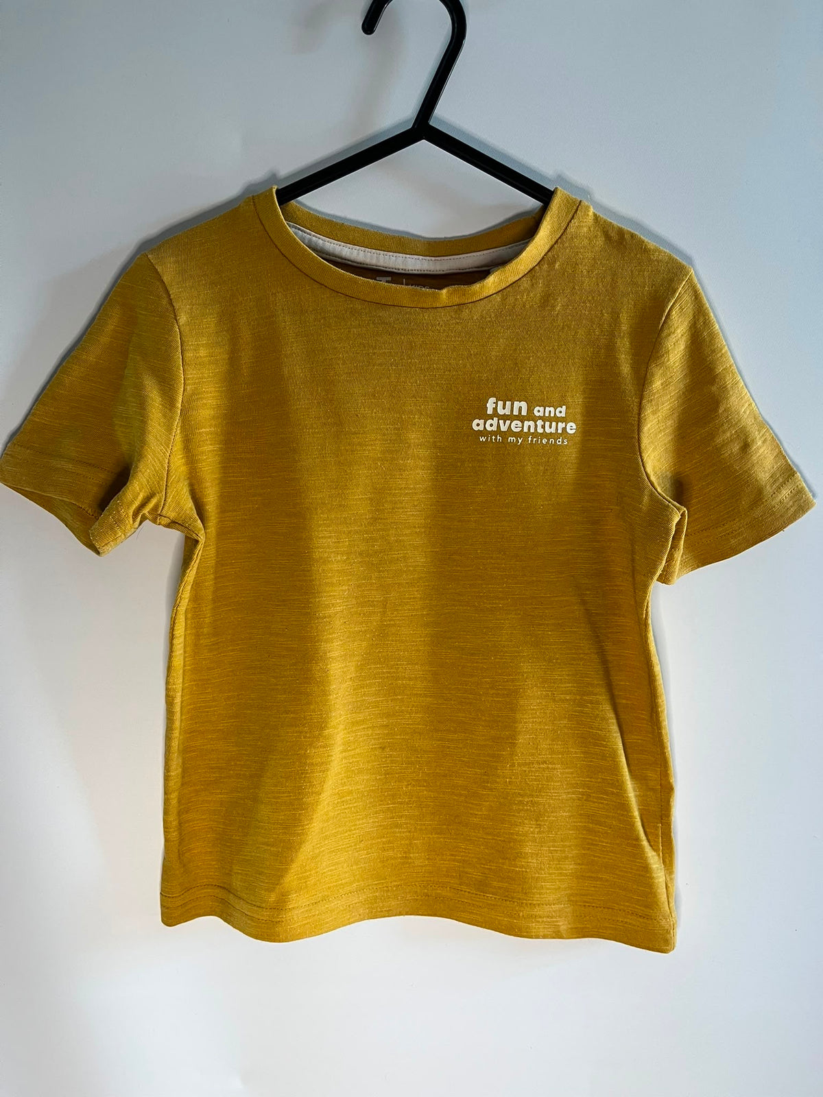 A mustard yellow short-sleeve t-shirt hanging on a black hanger against a plain white wall. The shirt features the text 'full and adventure with my friends' printed in white on the left chest. TU preloved little drums children clothing