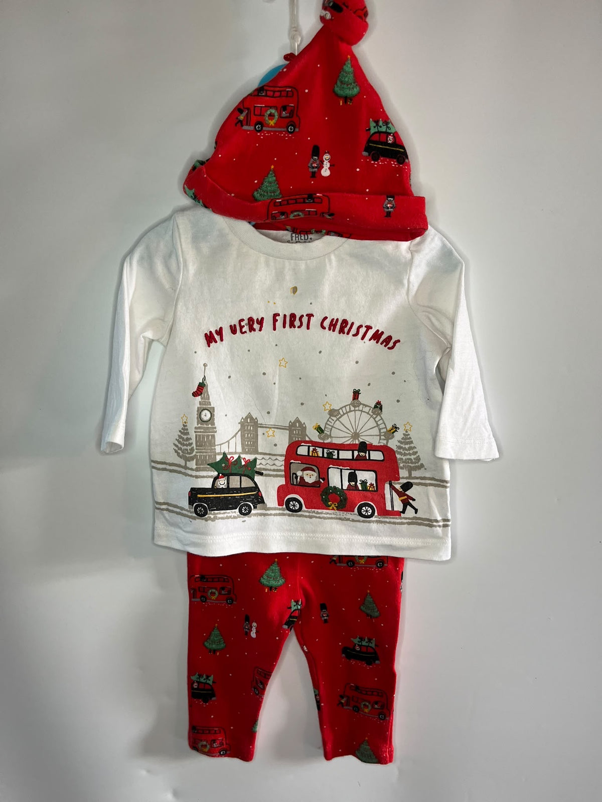 A baby Christmas outfit set hanging against a plain white wall, featuring a white long-sleeve top with a red and multicolor Christmas city graphic and the text 'MY VERY FIRST CHRISTMAS,' red leggings with a red Christmas print, and a red Santa hat with holiday motifs.