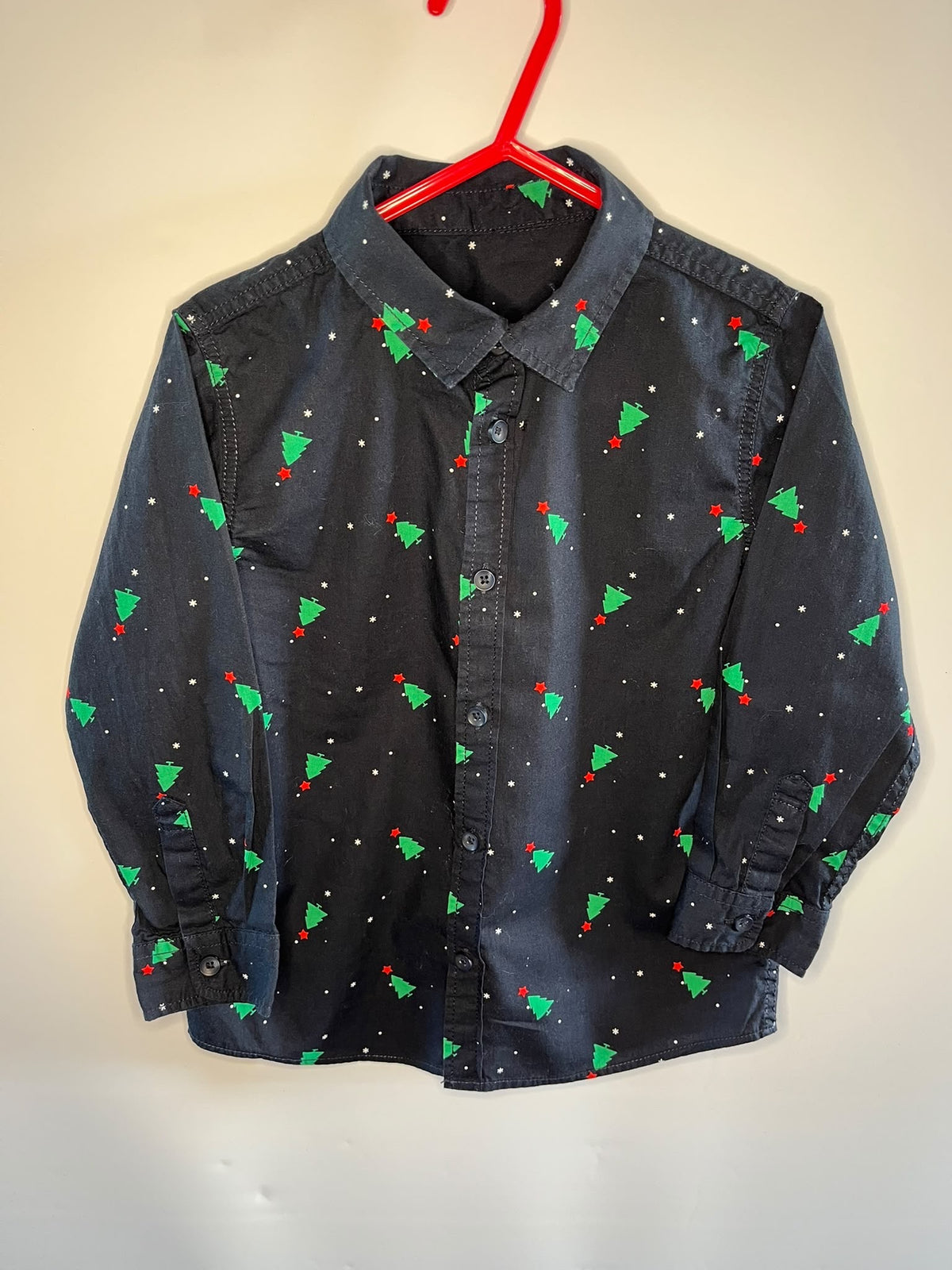 A long-sleeve button-up shirt for kids in a faded black or charcoal color, featuring a green Christmas tree and red berry print with small white dots, hanging on a red plastic hanger against a plain beige wall.