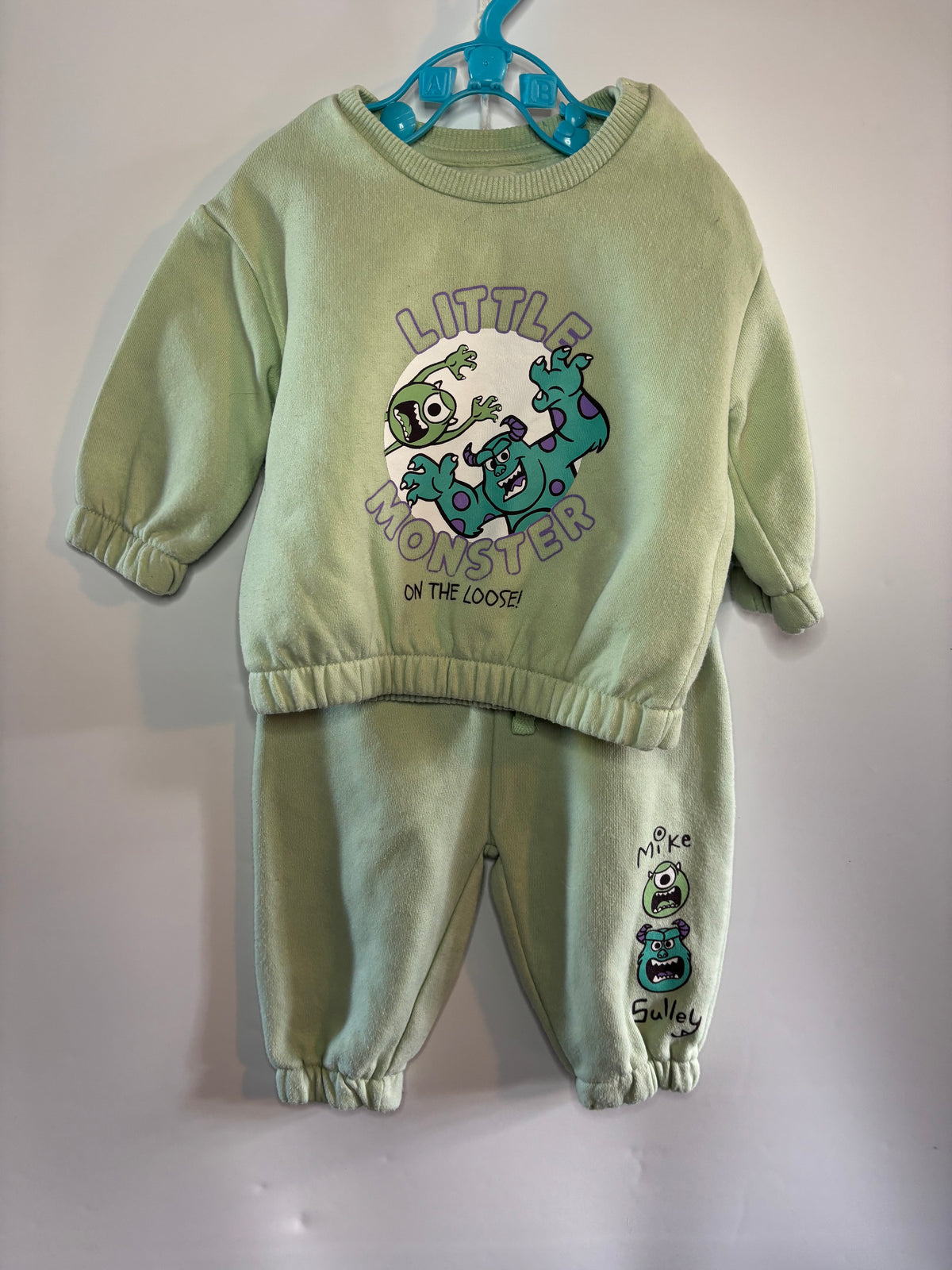 two-piece sweatshirt and jogger pants set in light sage green, featuring a cartoon monster graphic on the sweatshirt and pants, hanging on a blue hanger against a plain wall.