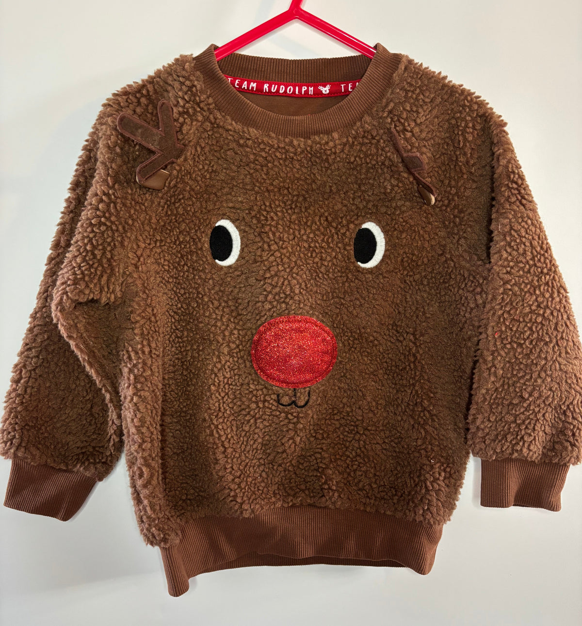 A brown sherpa fleece long-sleeve sweatshirt for kids, featuring a reindeer face design with embroidered black and white eyes and a red nose, hanging on a red hanger against a plain white wall.