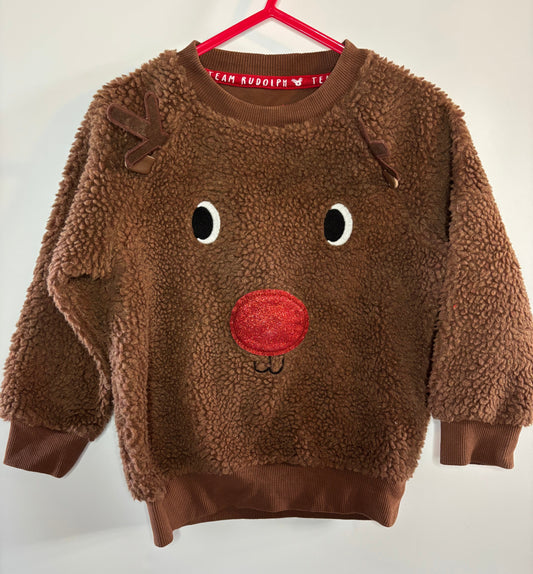 A brown sherpa fleece long-sleeve sweatshirt for kids, featuring a reindeer face design with embroidered black and white eyes and a red nose, hanging on a red hanger against a plain white wall.
