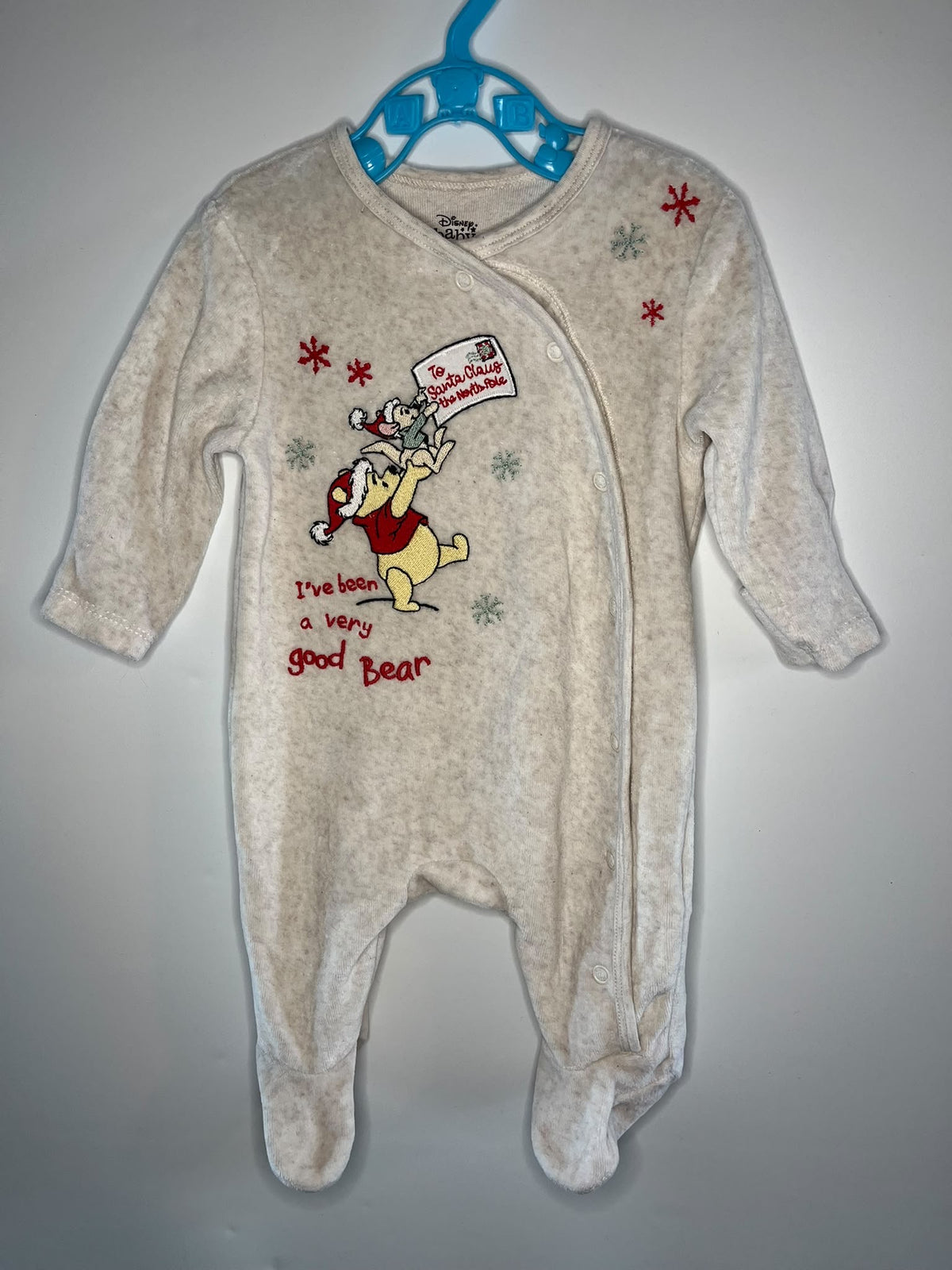 A baby footed sleeper in a heathered cream-beige fabric, The sleeper features a Winnie the Pooh graphic with the text 'I've got a very good Bear' and red and green snowflake accents.