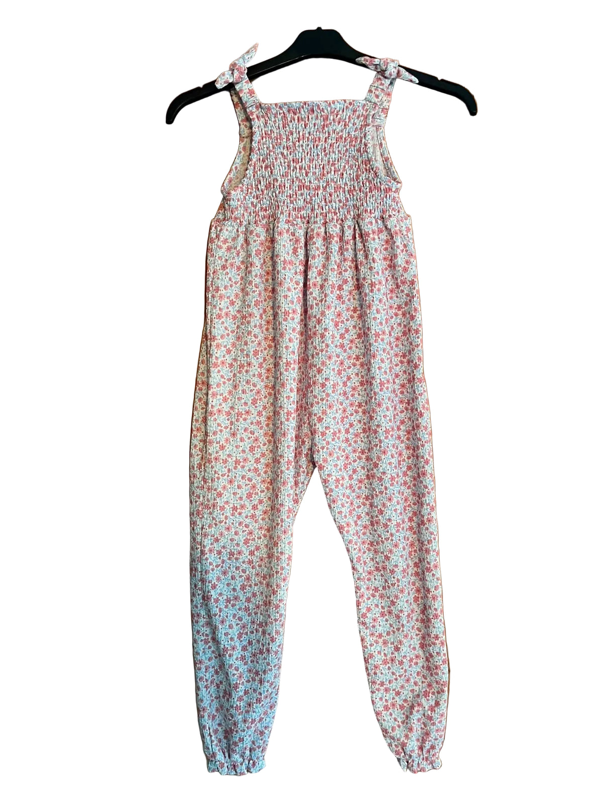 A children's sleeveless jumpsuit with a smocked bodice and tie shoulder straps, featuring a small pink and red floral print on a white background.