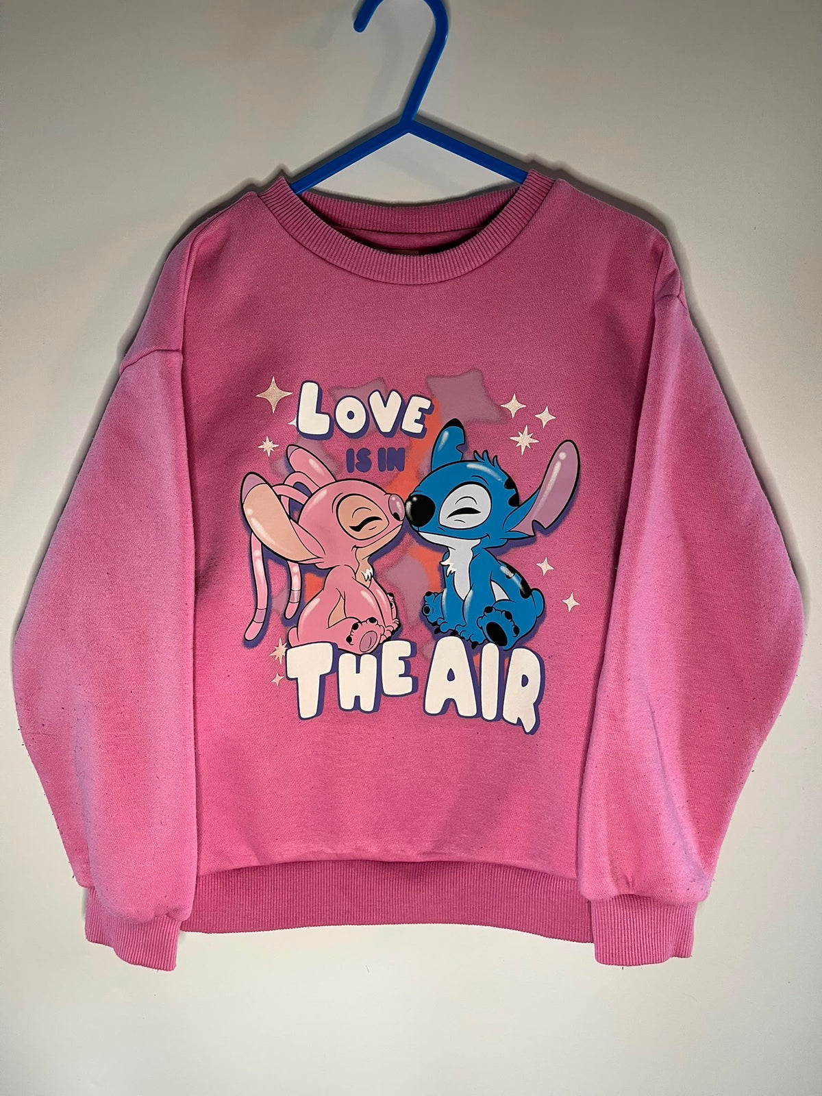 A pink crewneck sweatshirt hanging on a blue hanger against a plain light wall. The sweatshirt features a graphic print of Stitch and Angel with the text 'LOVE IS IN THE AIR'.