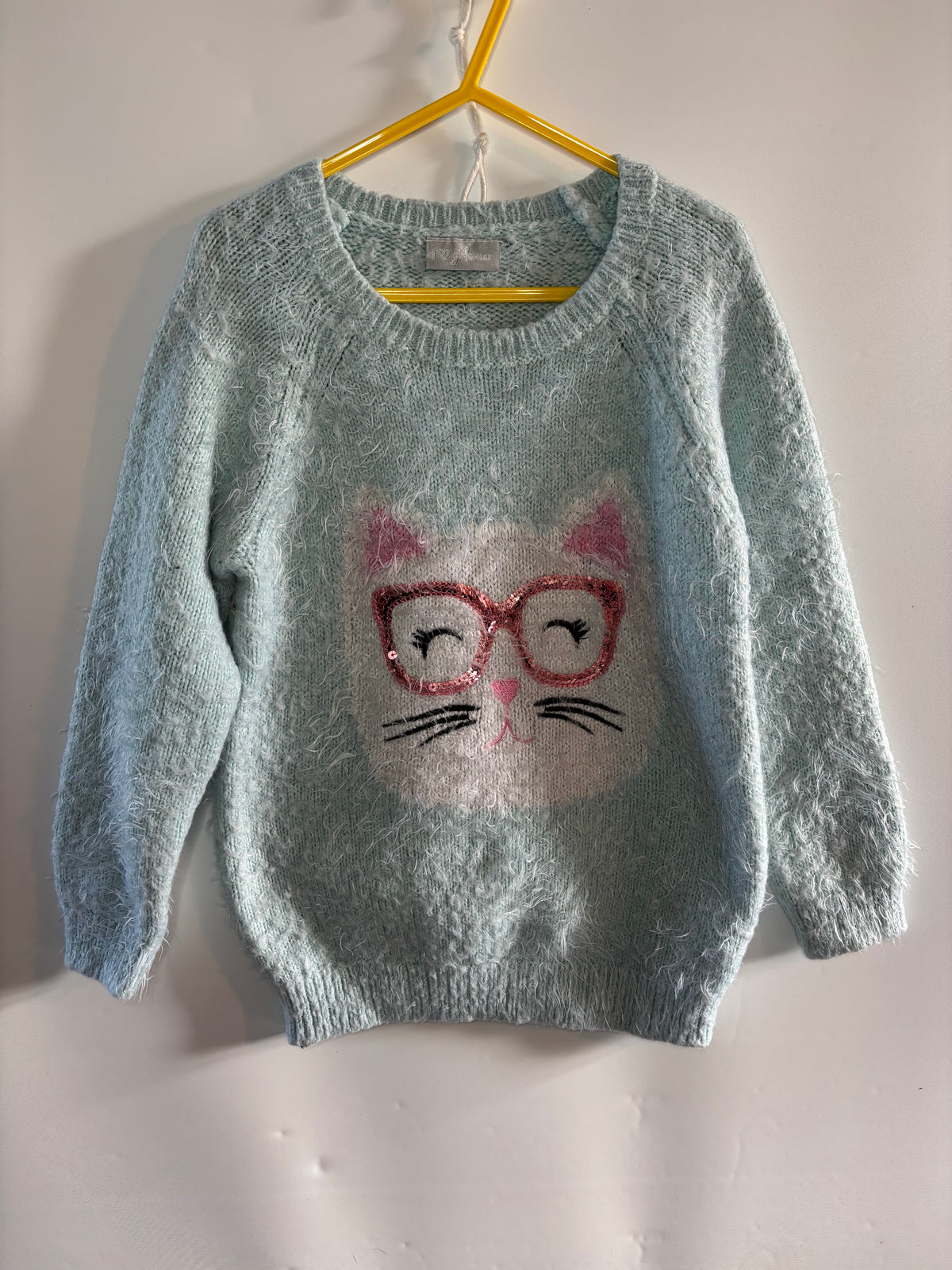 A light blue fuzzy knit sweater hanging on a yellow hanger against a white wall. The sweater features a white cat face graphic with pink ears, red-rimmed glasses, and black whiskers on the front.