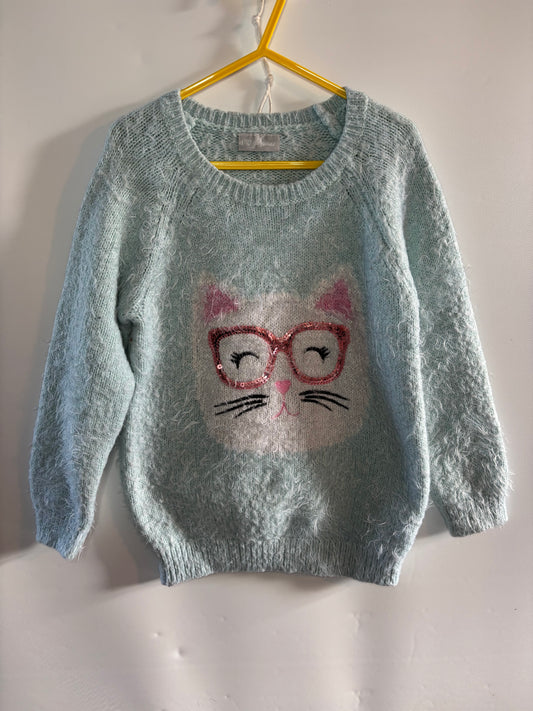A light blue fuzzy knit sweater hanging on a yellow hanger against a white wall. The sweater features a white cat face graphic with pink ears, red-rimmed glasses, and black whiskers on the front.
