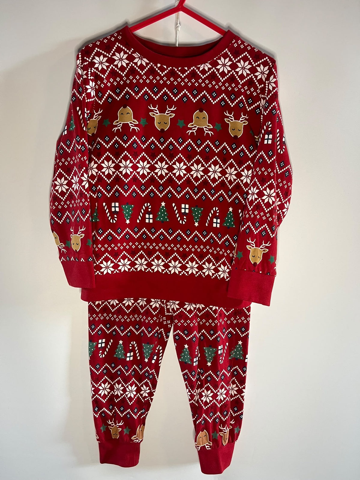 A red Christmas-themed matching sweatshirt and jogger pants set.. The long-sleeve top and pants feature white snowflake and geometric patterns, green pine trees, and gold reindeer motifs.