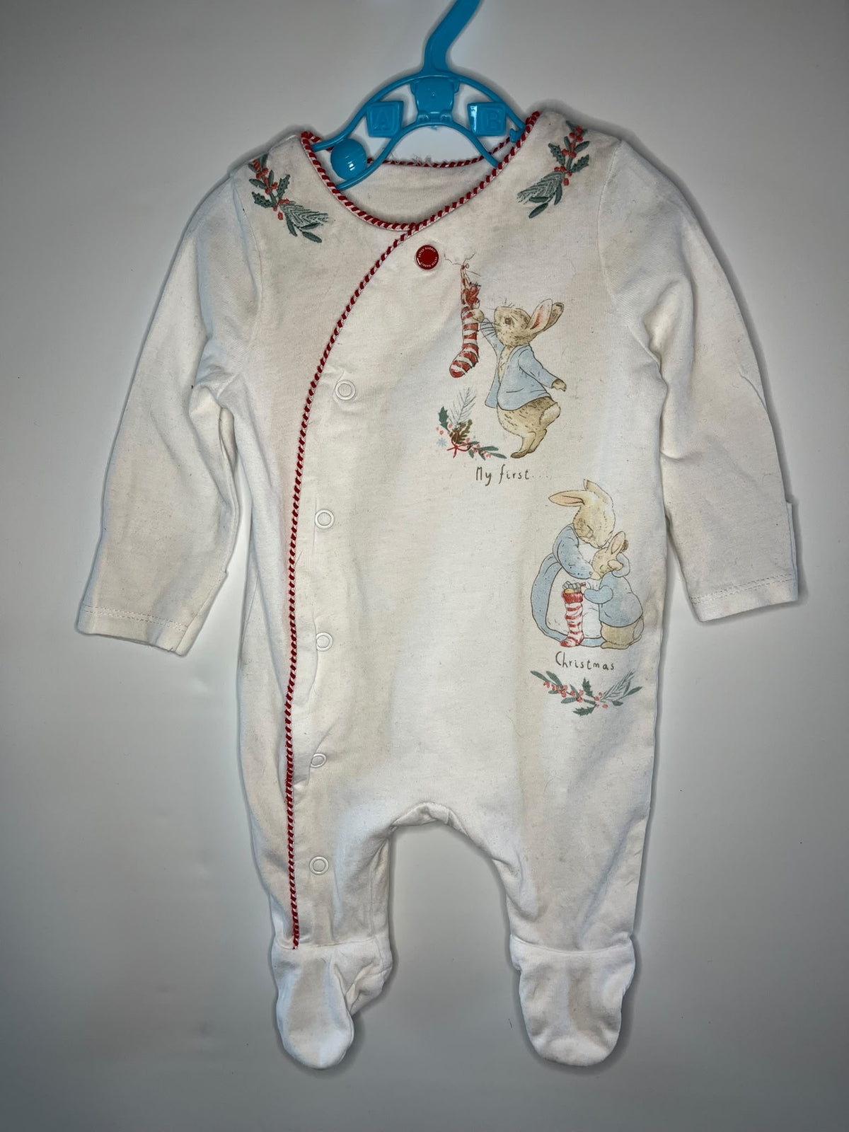 A baby footed sleeper in cream white, decorated with red stitching, a red snap button, and illustrations of Beatrix Potter's characters, hanging on a blue plastic hanger against a plain gray wall.