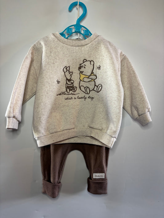 A children's two-piece outfit hanging on a blue hanger against a plain light gray wall. The set includes a cream-colored sweatshirt with a Winnie the Pooh graphic and text on the front, paired with dark brown pants.