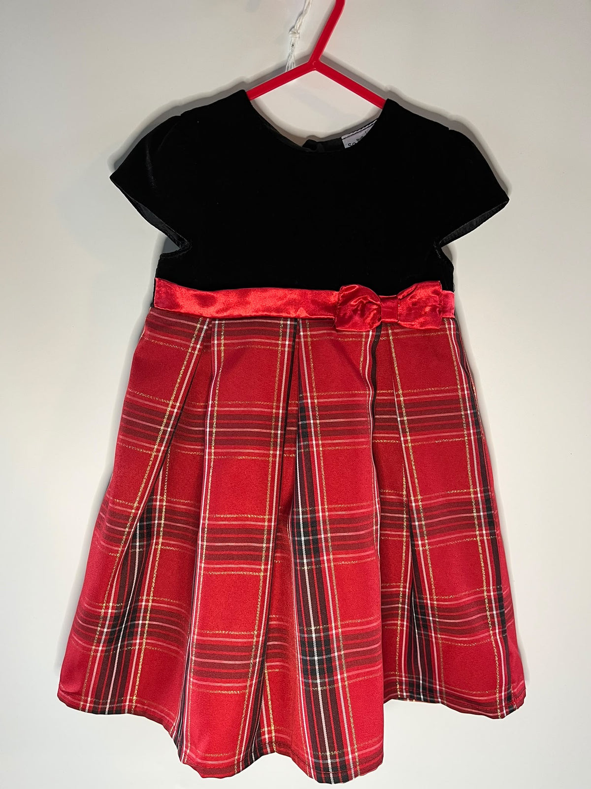 A toddler girl's dress. The dress features a solid black velvet short-sleeve bodice and a pleated red tartan plaid skirt with a matching red satin waistband and bow.