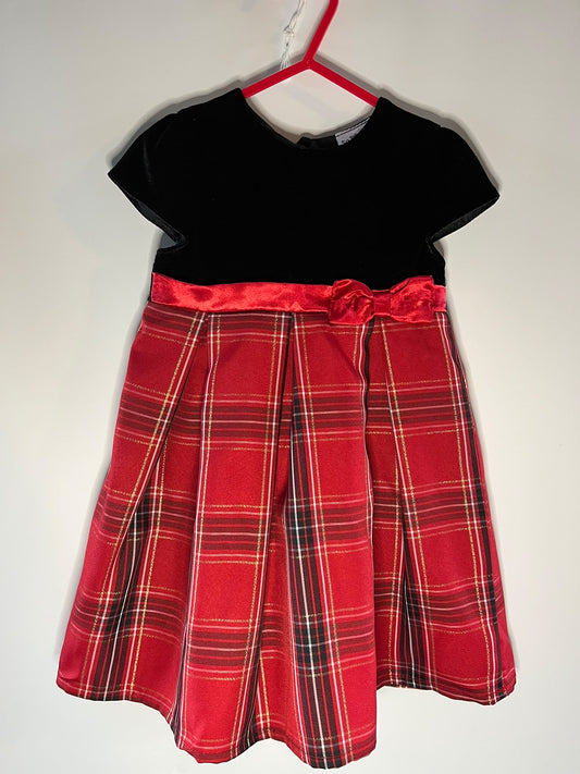 A toddler girl's dress. The dress features a solid black velvet short-sleeve bodice and a pleated red tartan plaid skirt with a matching red satin waistband and bow.