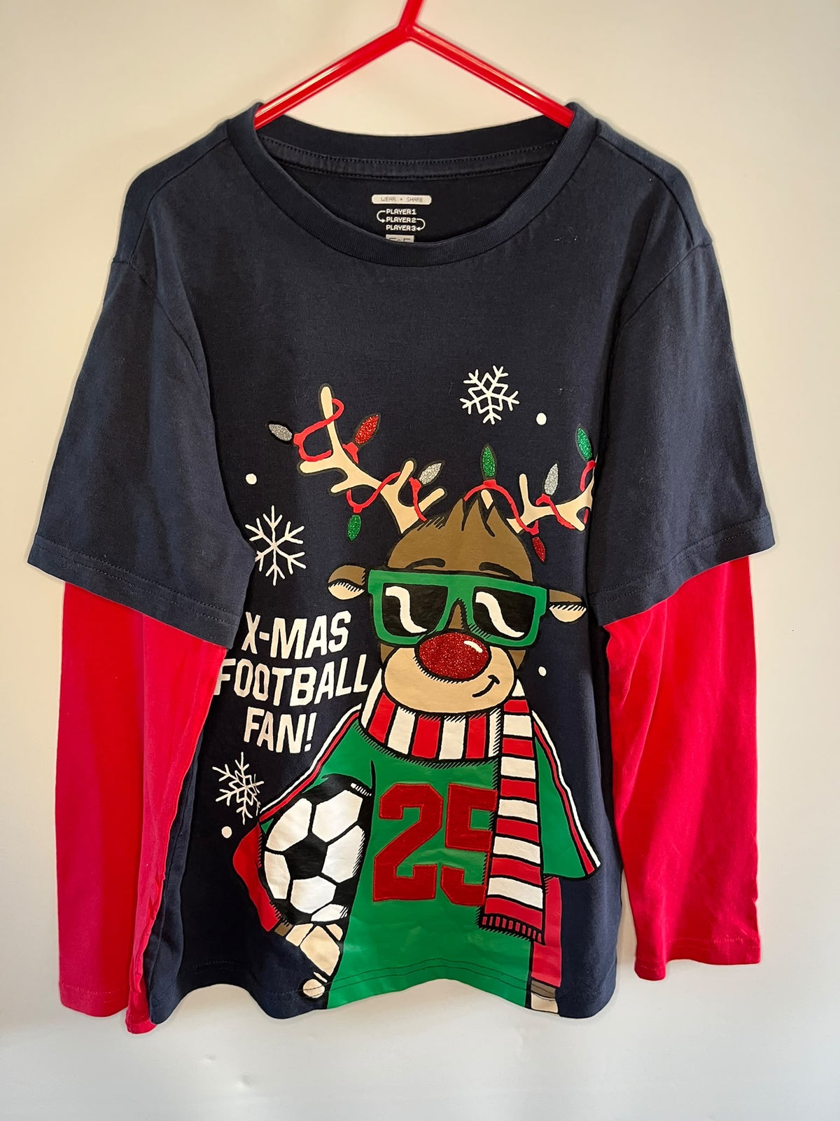 A navy blue long-sleeve t-shirt with bright red sleeves, featuring a festive Christmas football graphic of a reindeer wearing green sunglasses and a red and white striped scarf. The graphic includes the text 'X-MAS FOOTBALL FAN!' and the number '25'.