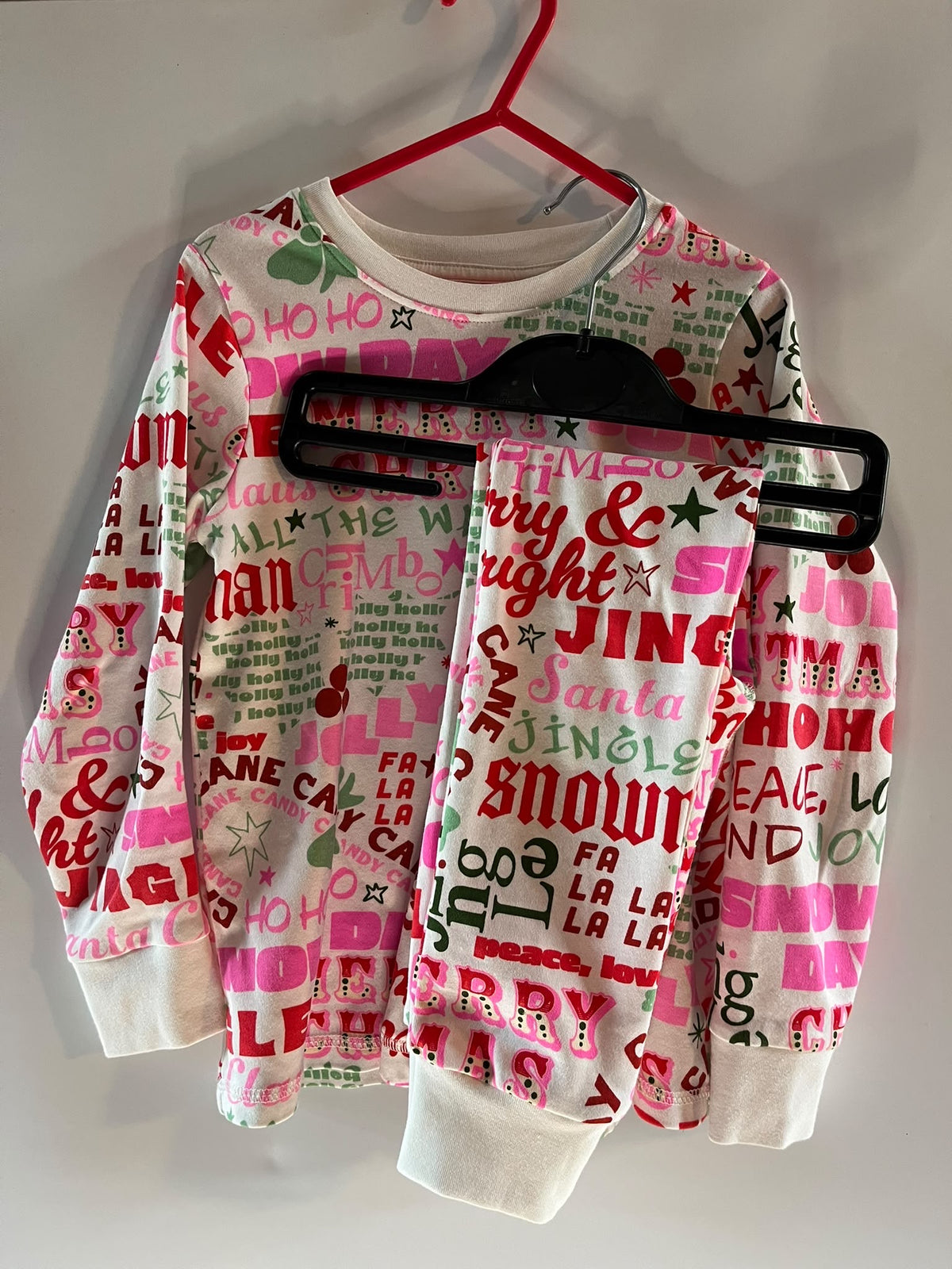 A children's long-sleeve sweatshirt and matching pants set hanging on a red hanger against a plain wall. The white fabric is covered in a colorful Christmas-themed typographic print with words like 'Merry & Bright', 'Santa', 'Jingle', 'Snow', and 'Ho Ho Ho' in pink, green, red, and black. Next preloved little drums 