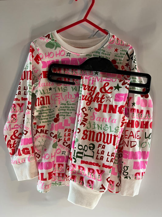 A children's long-sleeve sweatshirt and matching pants set hanging on a red hanger against a plain wall. The white fabric is covered in a colorful Christmas-themed typographic print with words like 'Merry & Bright', 'Santa', 'Jingle', 'Snow', and 'Ho Ho Ho' in pink, green, red, and black. Next preloved little drums 