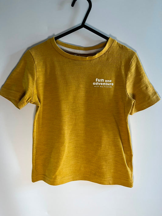 A mustard yellow short-sleeve t-shirt hanging on a black hanger against a plain white wall. The shirt features the text 'full and adventure with my friends' printed in white on the left chest. TU preloved little drums children clothing 