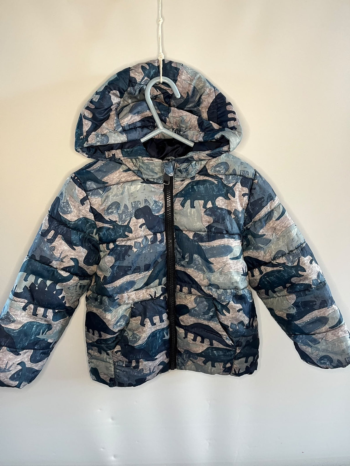 A hooded puffer jacket with a dinosaur camouflage pattern in shades of blue, gray, and beige, hanging on a white plastic hanger against a plain off-white wall.