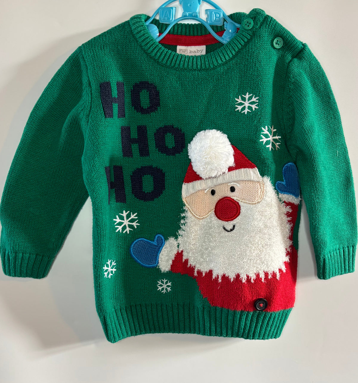 A green knit baby sweater featuring a festive Christmas design with 'HO HO HO' text, white snowflakes, and a plush embroidered Santa Claus face, red suit, and blue gloves.