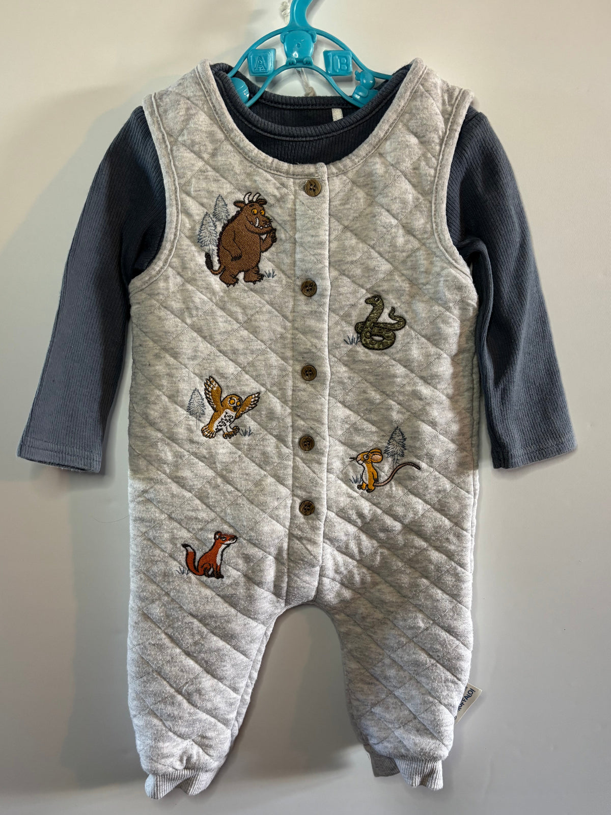 A baby's quilted footless vest romper in a light heather gray, featuring embroidered woodland animals (bear, fox, duck, squirrel) and a snap-button front, layered over a dark gray long-sleeve shirt and hanging on a blue hanger against a plain wall.