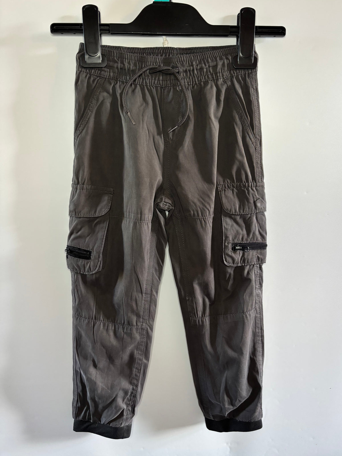 Dark gray cargo jogger pants hanging on a black hanger against a plain white wall. The pants feature an elastic waistband, drawstring, side cargo pockets with zippered details, and elastic cuffs.