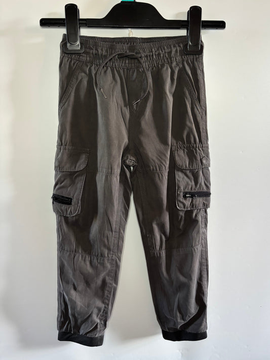Dark gray cargo jogger pants hanging on a black hanger against a plain white wall. The pants feature an elastic waistband, drawstring, side cargo pockets with zippered details, and elastic cuffs.