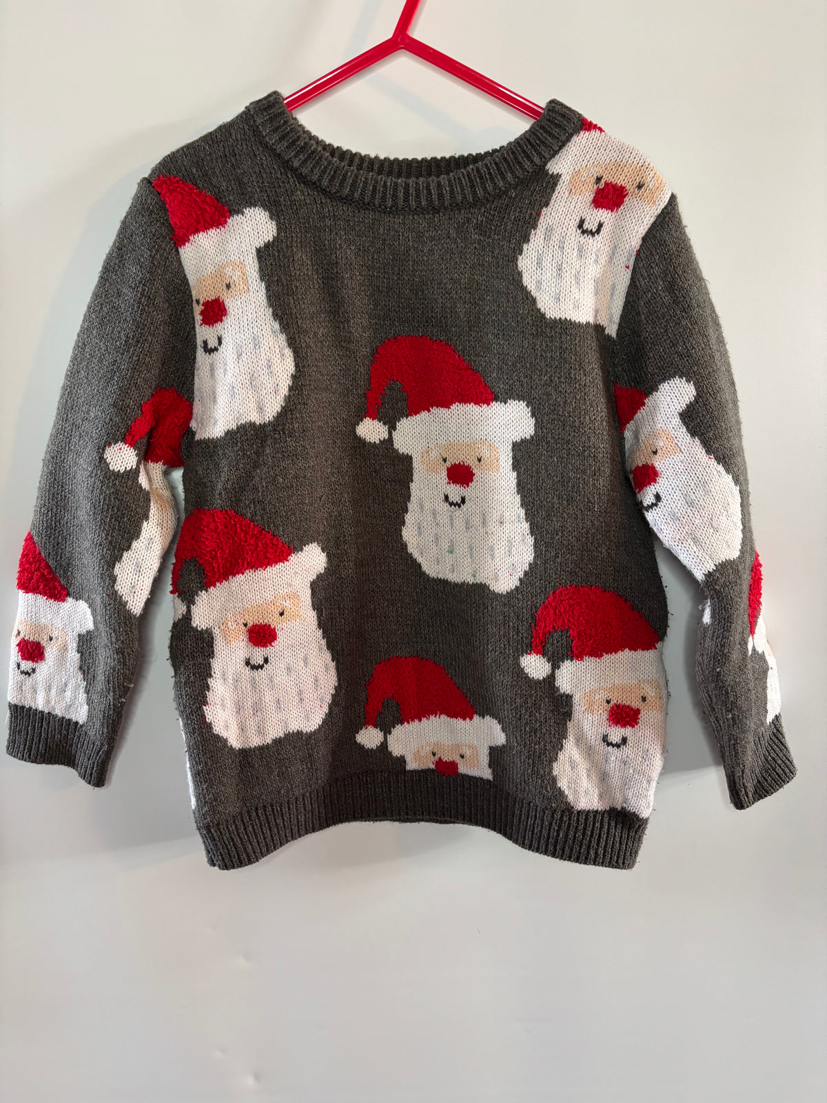 A dark gray knit Christmas sweater featuring a repeating pattern of Santa Claus faces in red and white, hanging on a red hanger against a plain light gray wall.