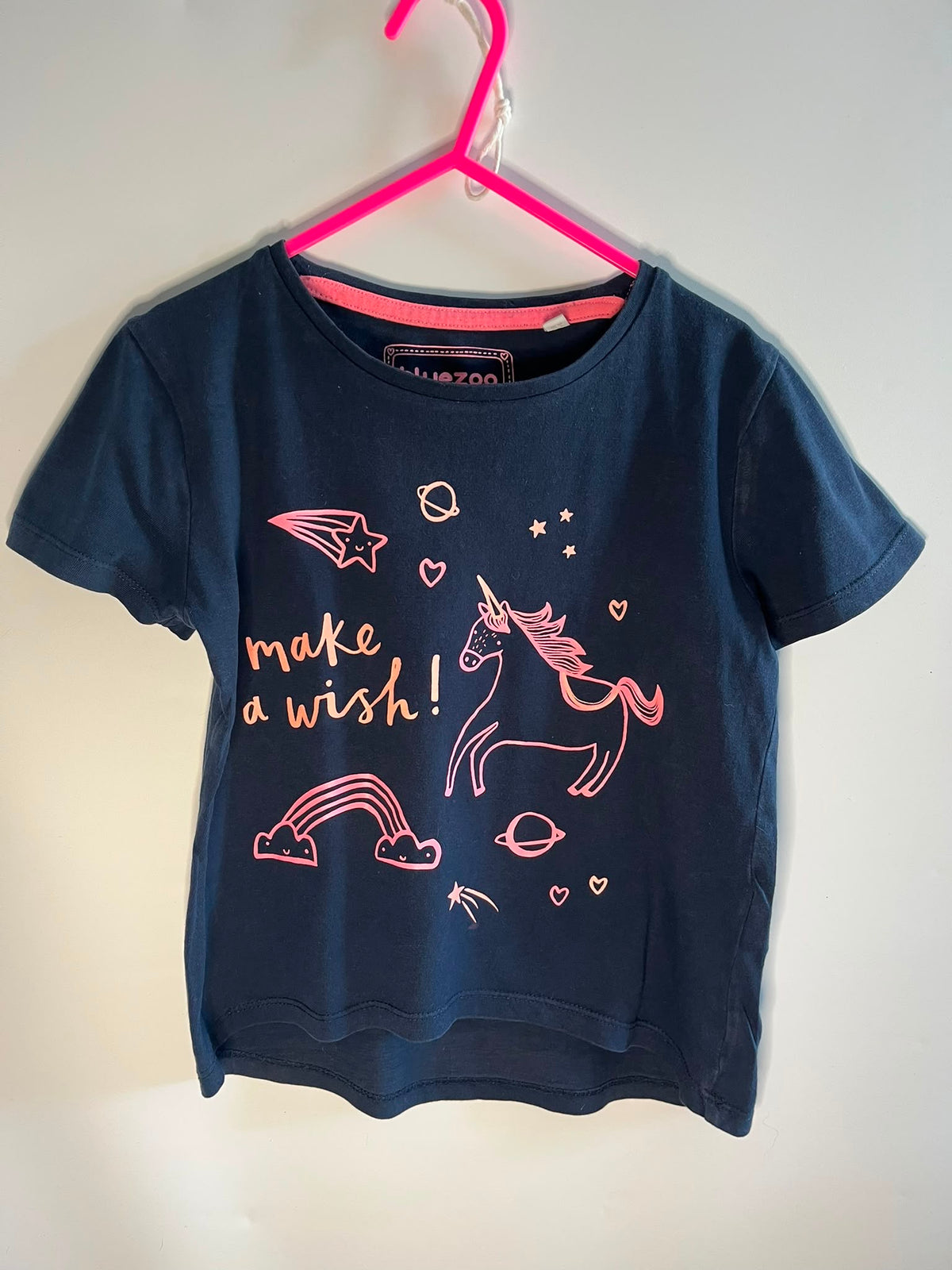 A navy blue short-sleeve t-shirt with a pink unicorn and 'make a wish!' graphic, hanging on a bright pink hanger against a plain light wall.