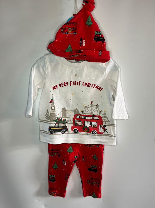 A baby Christmas outfit set hanging against a plain white wall, featuring a white long-sleeve top with a red and multicolor Christmas city graphic and the text 'MY VERY FIRST CHRISTMAS,' red leggings with a red Christmas print, and a red Santa hat with holiday motifs.