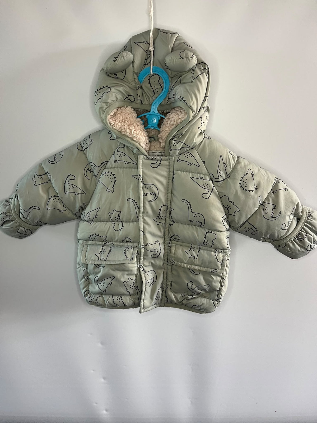 A light sage green hooded puffer jacket with a dinosaur print pattern, featuring a beige sherpa lining inside the hood,