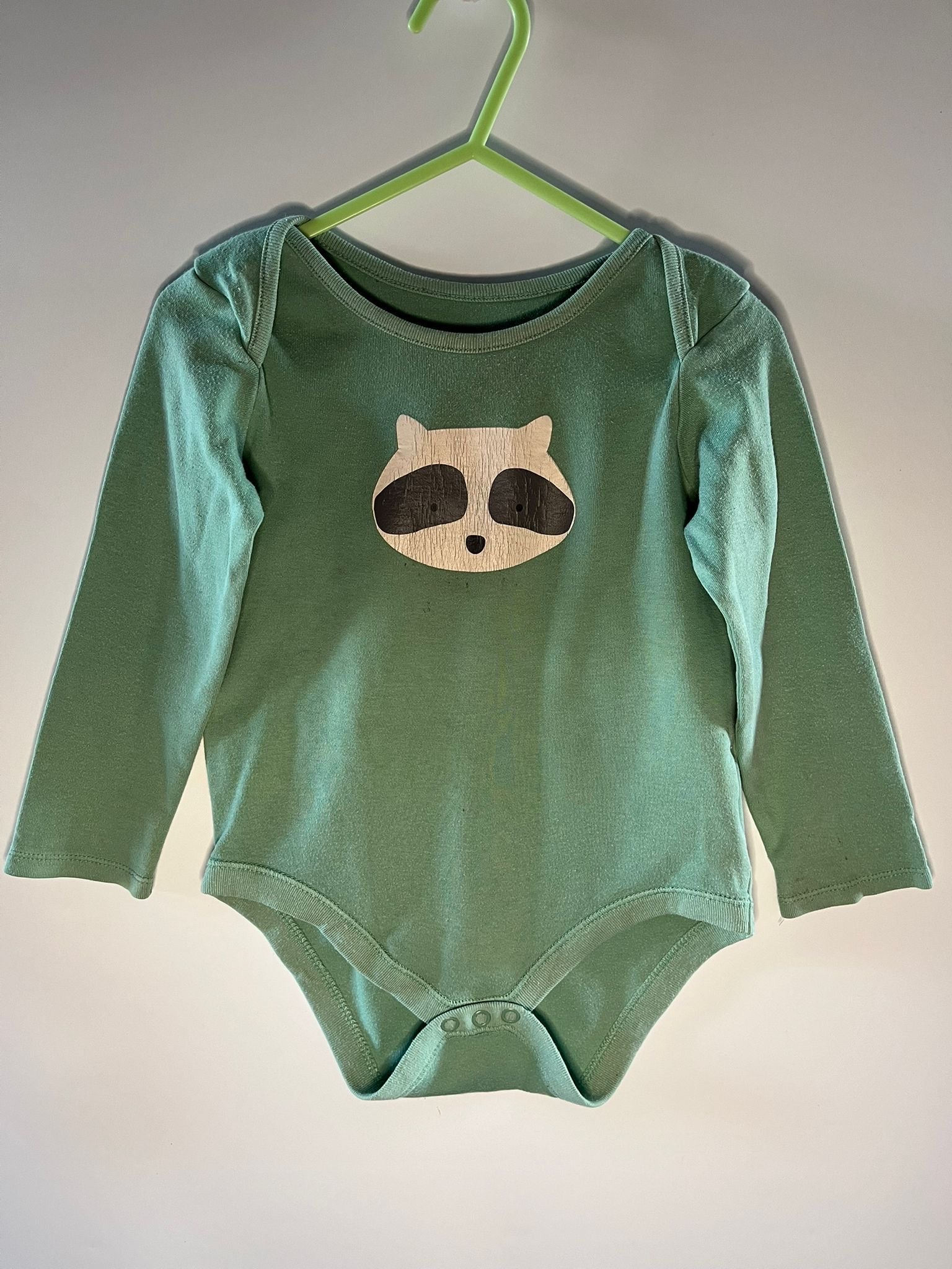 Green baby onesie with a raccoon design on a hanger against a plain background