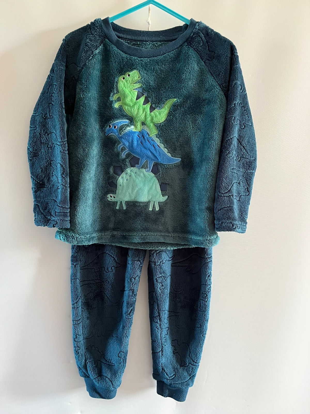 A two-piece children's pajama set hanging on a turquoise hanger against a plain white wall. The set includes a long-sleeve top and matching pants in a dark teal blue plush fleece fabric with a dinosaur pattern, featuring a graphic of three colorful dinosaurs (green, blue, and light green) on the front.