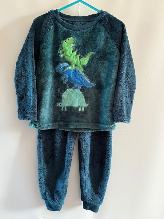A two-piece children's pajama set hanging on a turquoise hanger against a plain white wall. The set includes a long-sleeve top and matching pants in a dark teal blue plush fleece fabric with a dinosaur pattern, featuring a graphic of three colorful dinosaurs (green, blue, and light green) on the front.